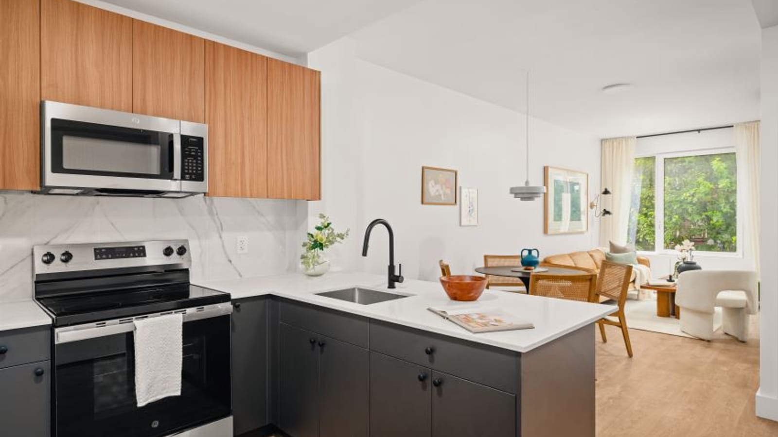 Kitchen with dark lower cabinets, wood upper cabinets, a stainless steel range and microwave, opening onto a living room with a window