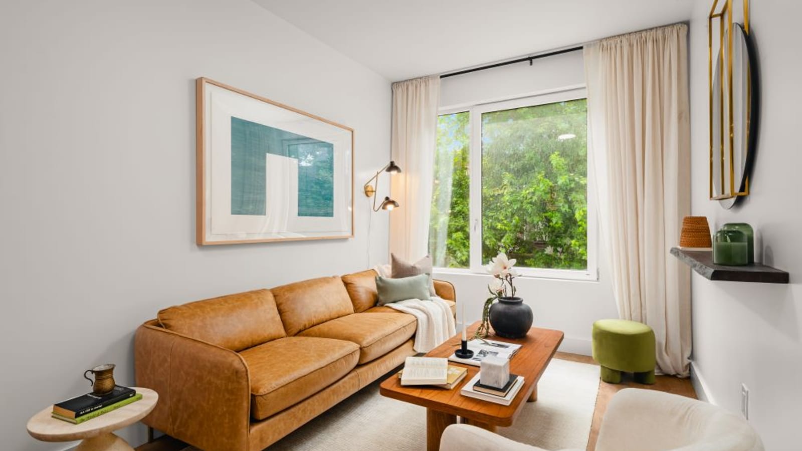 Living room with a tan leather sofa, a round wooden coffee table and a wide window with curtains