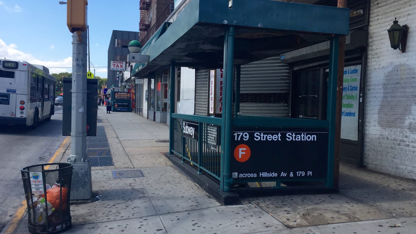 A green subway entrance canopy with a black sign reading 179 Street Station, an orange F line symbol and the words across Hillside Av & 179 Pl with a wheelchair symbol, on a wide sidewalk beside shuttered storefronts; a city bus is at the curb at left
