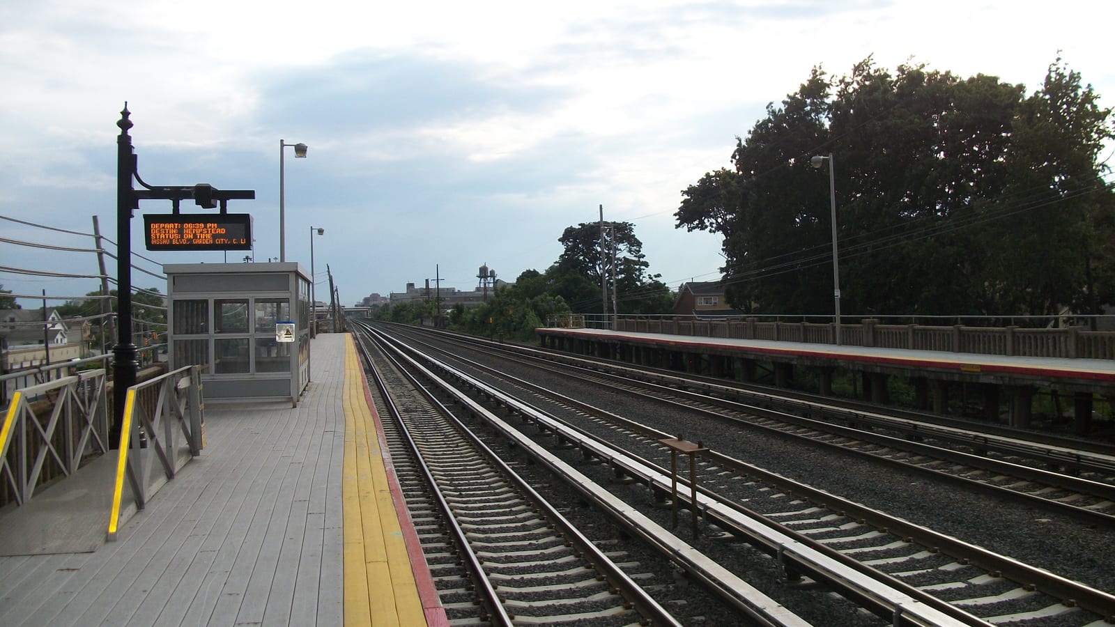 A raised railroad platform with gray wooden boards, a yellow edge strip, a glass shelter and an orange departure sign, beside four sets of tracks and a second platform with a stone railing under a cloudy sky