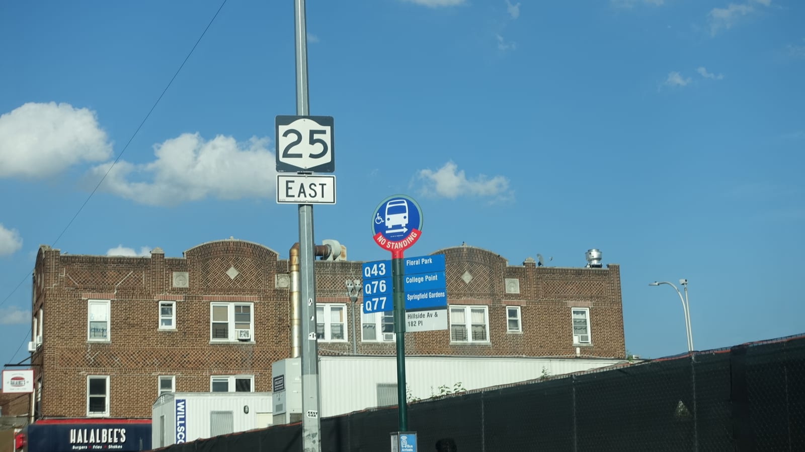 A tall pole with a white New York Route 25 East sign above a round blue bus stop sign reading No Standing, with route plates for Q43, Q76 and Q77 and a plate reading Hillside Av & 182 Pl, in front of a three-story brick building with patterned brickwork under a blue sky; a black construction fence runs along the right