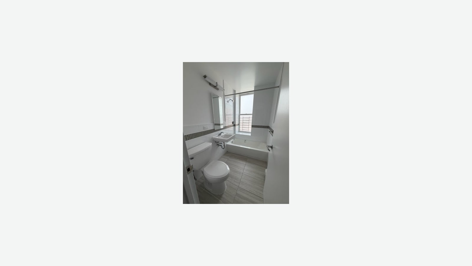 Photograph of a small white-tiled bathroom with a band of gray mosaic tile, a wall sink with exposed pipes under a mirror and light bar, a toilet, a bathtub with a shower rod, gray floor tiles and a barred window over the tub, shown small in the middle of a plain light background