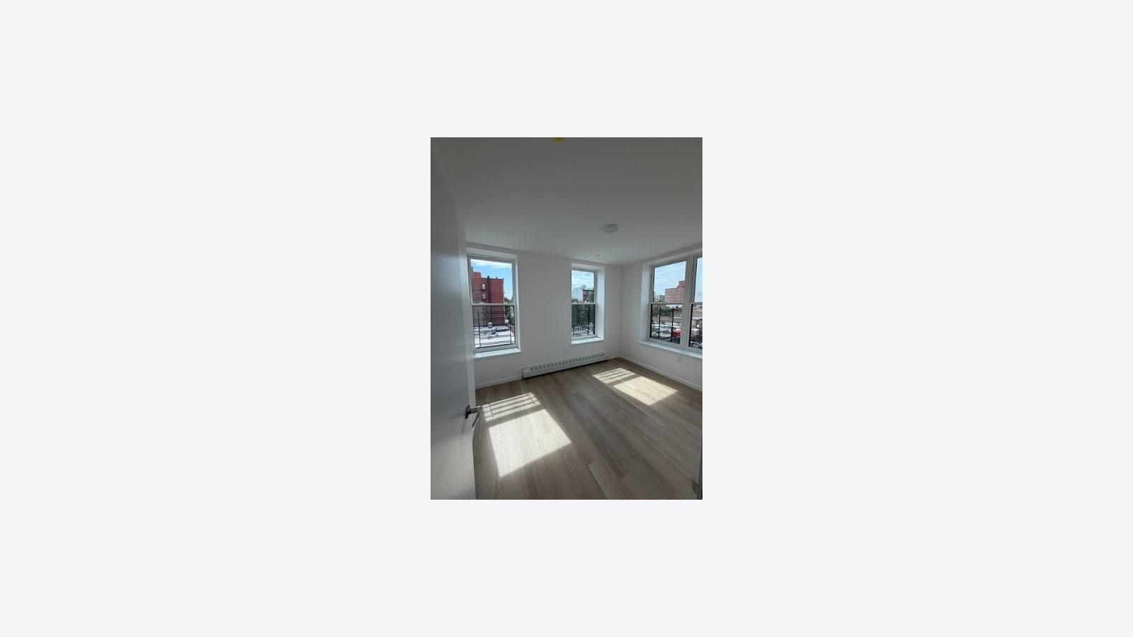 Photograph of an empty white bedroom with light wood-look floors, sunlight on the floor, a baseboard heater and three windows with railings looking out at brick buildings, shown small in the middle of a plain light background