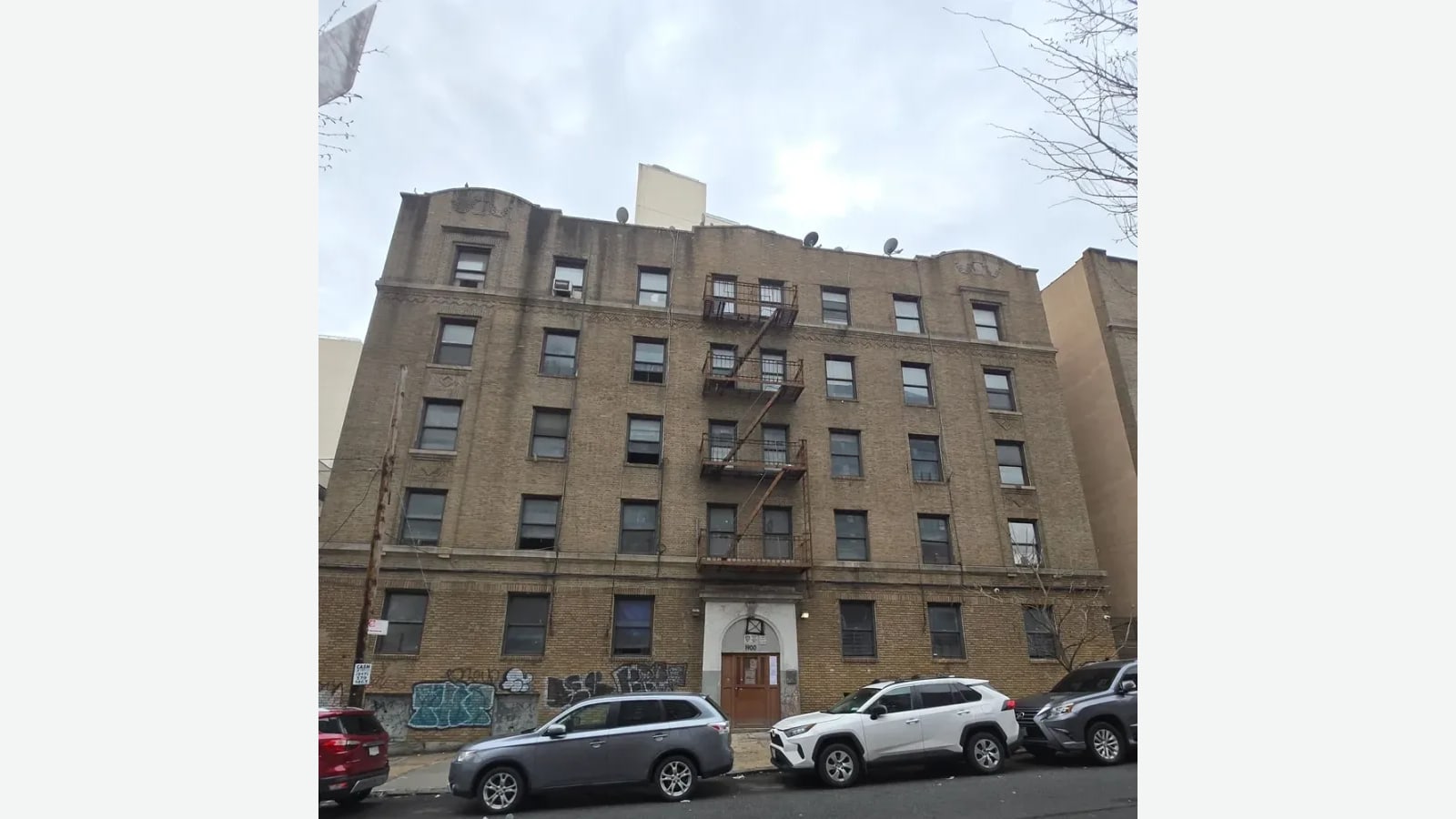 Photograph of the same style of tan brick apartment building under a gray sky before the work, with a rusted fire escape, older windows, satellite dishes on the roof, graffiti along the ground floor, a wooden door under a stone arch with the number 1900 above it, and parked cars along the curb