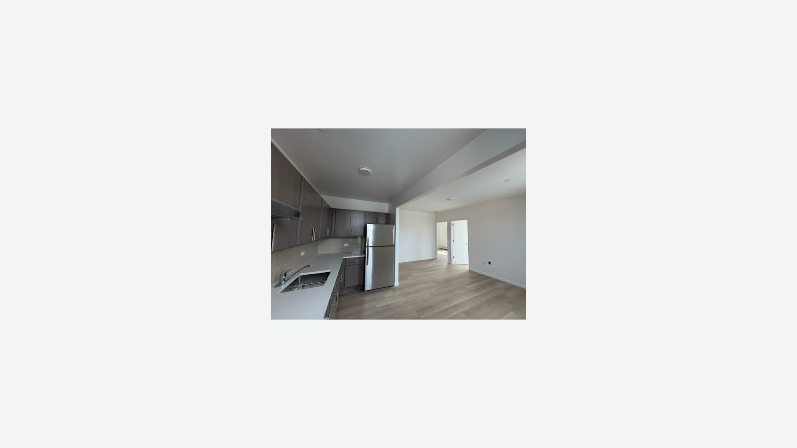 Photograph of an empty open kitchen with dark wood-look cabinets, a light gray counter with a steel sink, a steel refrigerator and a range hood, opening onto a white living room with light wood-look floors and two doors at the back, shown small in the middle of a plain light background