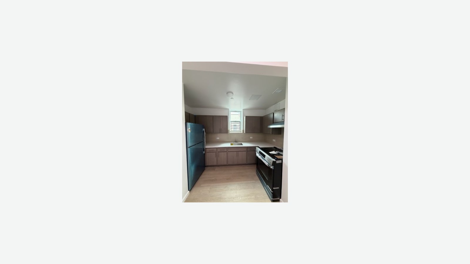 Photograph of an empty galley kitchen with dark wood-look cabinets, a pale tile backsplash, a white counter with a sink under a small barred window, a dark refrigerator at left, a range with a hood at right and light wood-look floors, shown small in the middle of a plain light background