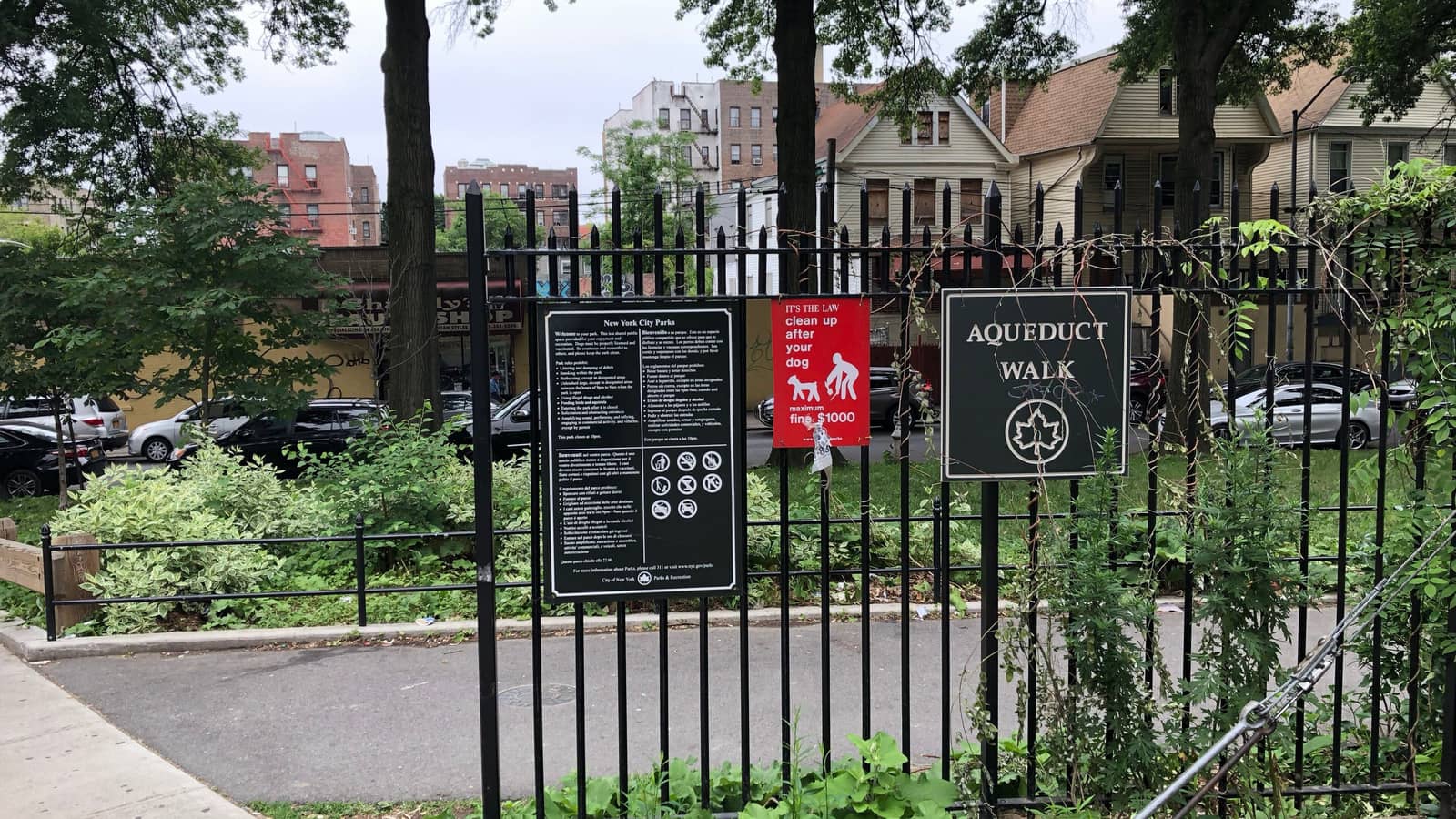 Photograph of a black iron park fence with a green sign reading Aqueduct Walk with a leaf logo, a New York City Parks rules sign and a red dog-waste sign, a paved path and shrubs behind it, and frame houses and brick apartment buildings across the street