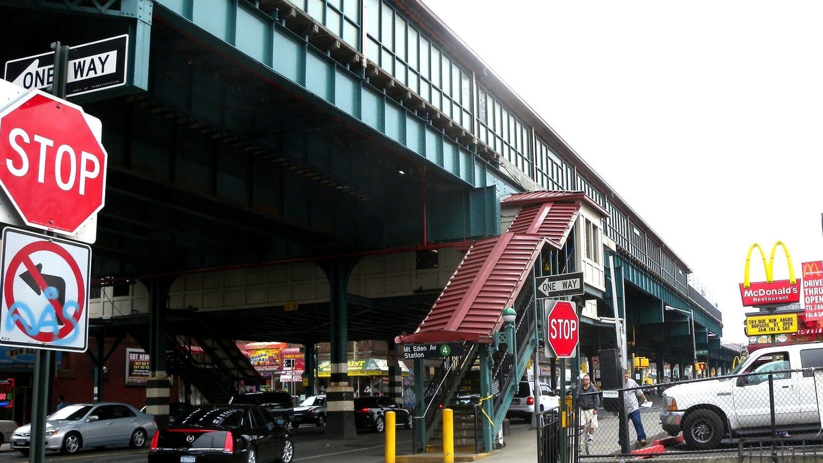 Photograph of a green elevated subway structure over a street, with a red-roofed staircase leading up to a sign reading Mt Eden Av Station and a 4 symbol, stop and one-way signs at left, parked cars, two people on the sidewalk and a fast-food sign at right