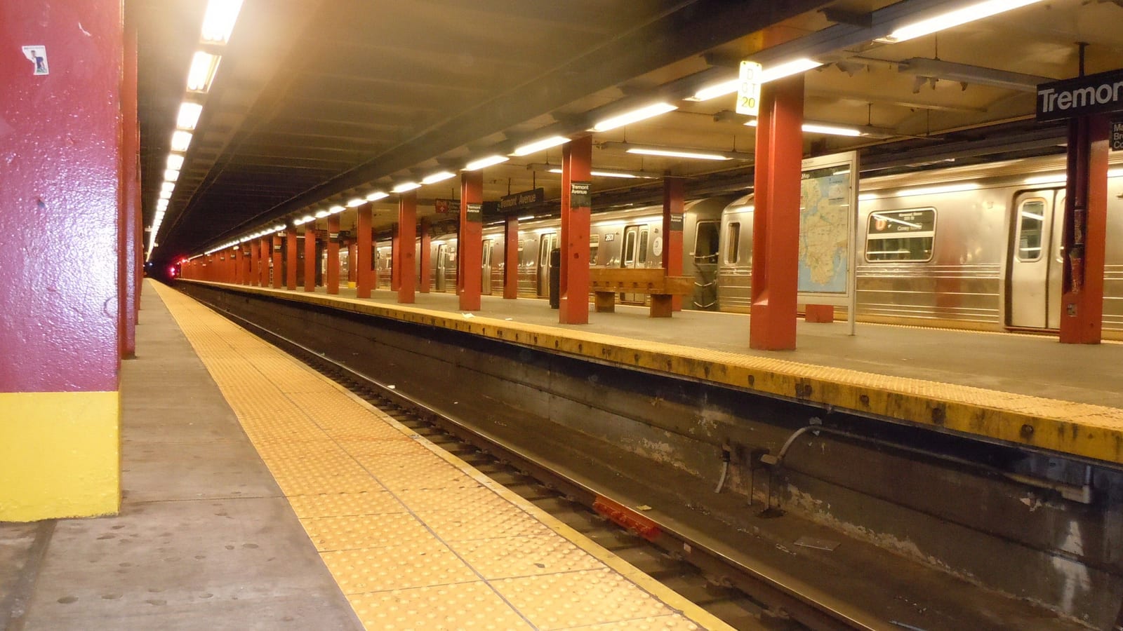 Photograph of an underground subway platform with red steel columns, a yellow textured platform edge, a wooden bench, a subway map and a silver train stopped at the far platform, with a sign reading Tremont at top right