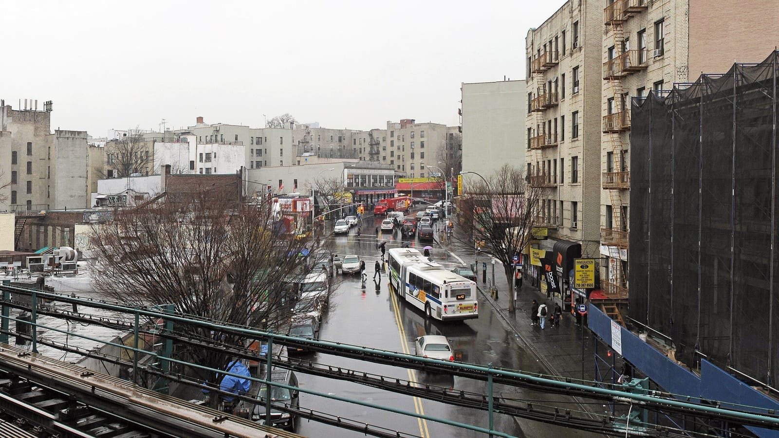Photograph from a raised subway platform on a gray, rainy day, looking down a wet avenue lined with five- and six-story apartment buildings, shops and bare trees, with a city bus, parked cars and a few people with umbrellas, green railings and track in the foreground, and a building wrapped in black scaffolding netting at right