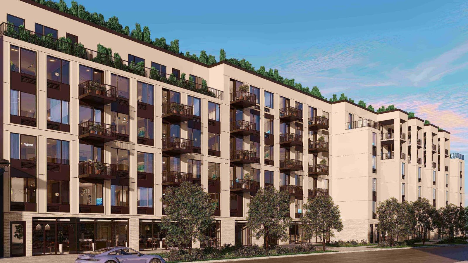 Rendering of a long six-story building with cream-colored panels, dark window frames and small dark balconies with potted plants, a row of trees along a roof terrace, glass storefronts with tables and chairs on the ground floor, young street trees and shrubs along the sidewalk, a lilac sports car parked at the curb at left, and a lower wing with stepped balconies at right, under a blue evening sky