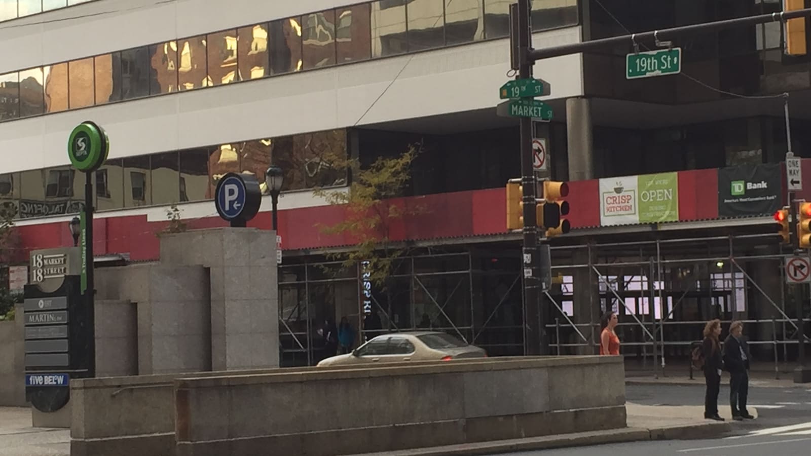 Street corner with a stone-walled stairway entrance marked by a green SEPTA sign, street signs for 19th Street and Market Street, scaffolding on the building behind and people waiting to cross