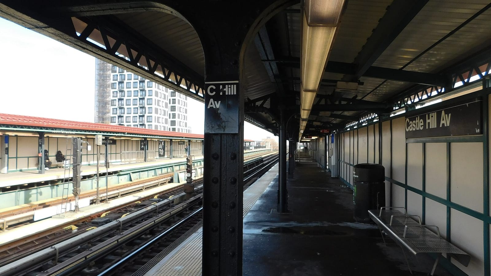 Photograph of a covered raised subway platform with a sign reading Castle Hill Av, green steel columns, a bench and a trash can, the tracks on the left and the far platform and a tall white apartment tower beyond