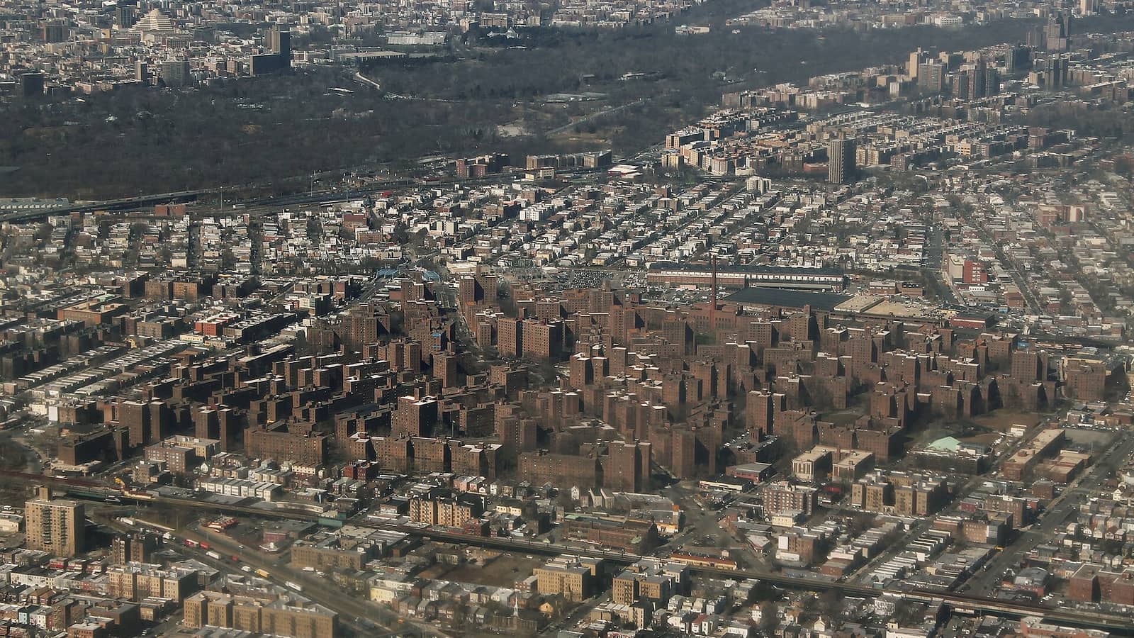 Aerial photograph of a dense part of the Bronx, with a large group of brown brick apartment towers in the middle, rows of smaller buildings around it, highways and a wooded park in the distance