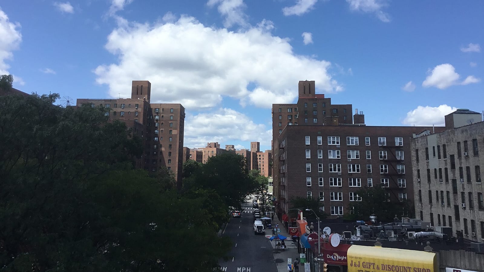 Photograph taken from a raised train platform of a street lined with tall brown brick apartment buildings, leafy trees on the left, parked cars and shop signs at street level, under a blue sky with white clouds