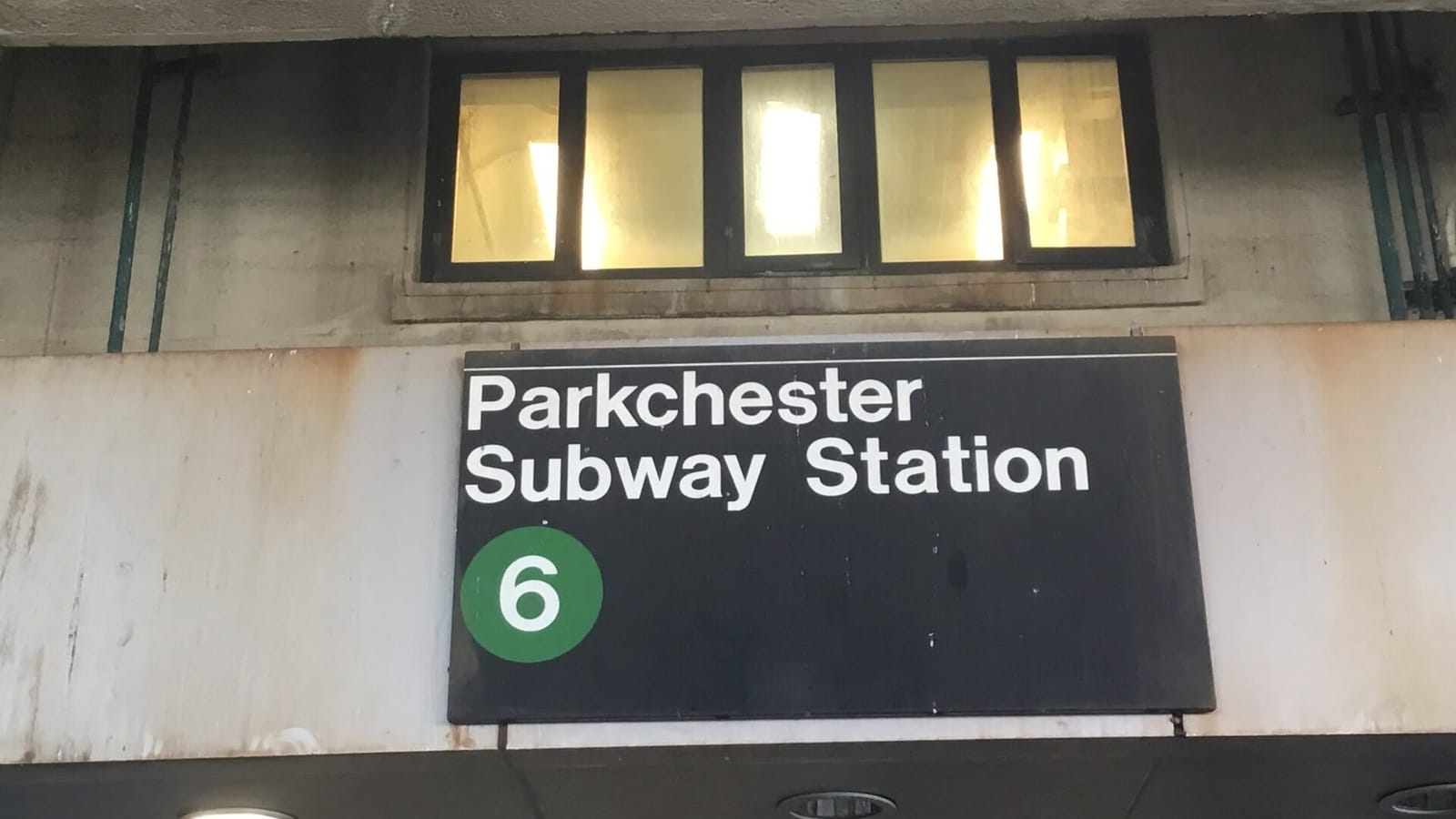 Photograph of a black sign reading Parkchester Subway Station with a green circle marked 6, on a weathered concrete wall below a row of lit windows