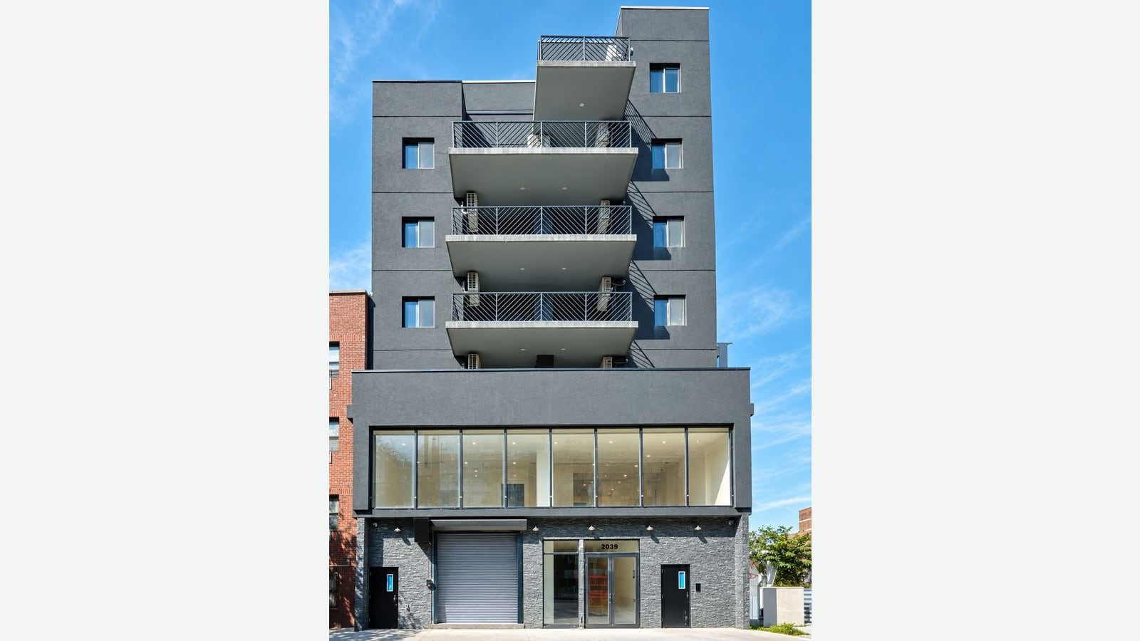 Photograph of a new mid-rise building with a dark gray stucco facade, a column of balconies with diagonal black railings in the middle, small square windows on each side, a wide glass-fronted second floor, and a ground floor of dark stone with a roll-down garage door, glass entrance doors under the number 2039 and two side doors, under a clear blue sky, shown on a plain light background