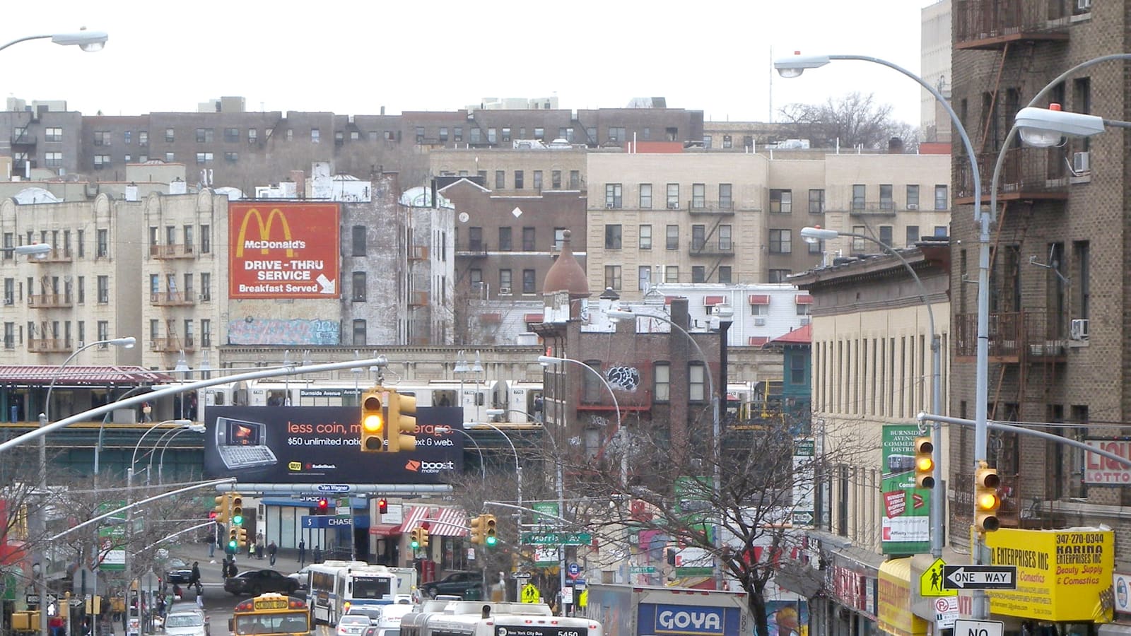 Photograph looking west along a busy street of shops and apartment buildings toward an elevated subway station, with buses, a yellow school bus, traffic lights and a large restaurant sign painted on a wall