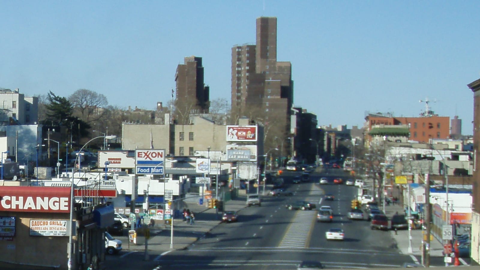 Photograph looking north along a wide avenue with traffic, a gas station sign, an oil change shop and low commercial buildings, with two tall brown brick apartment towers in the distance under a blue sky