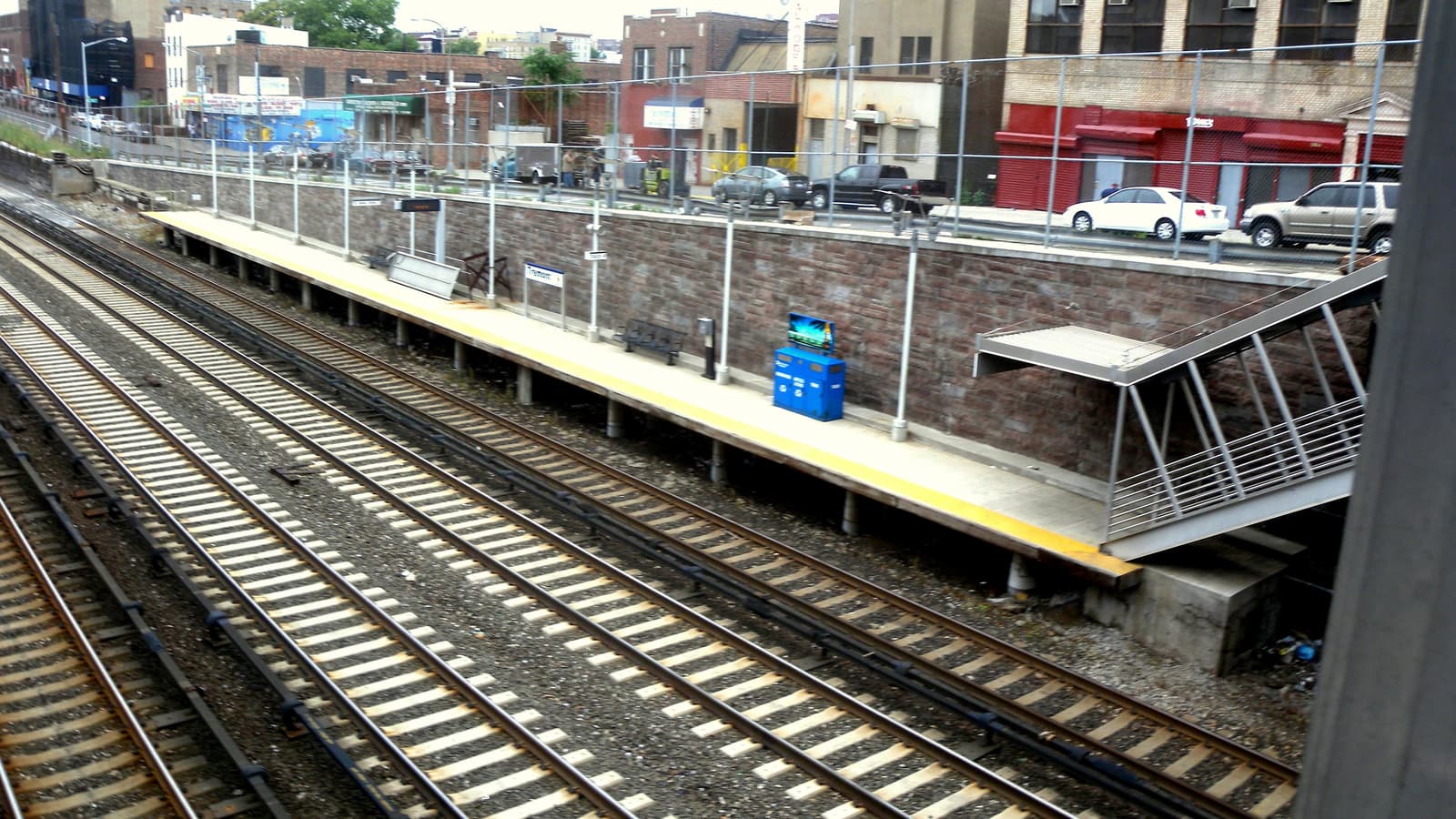Photograph of an open-air railroad platform with a yellow edge, a blue bin and a metal stair, below a stone retaining wall and a fence, with parked cars and low commercial buildings on the street above