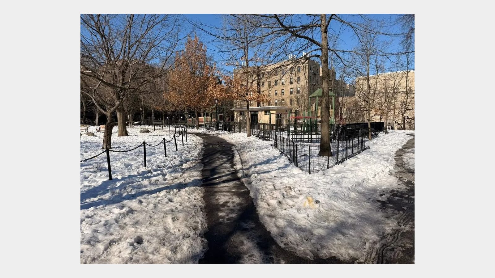 Photograph of a cleared park path through snow between bare trees, with a fenced playground with green roofs, a small park building and tall tan apartment buildings behind, under a blue sky