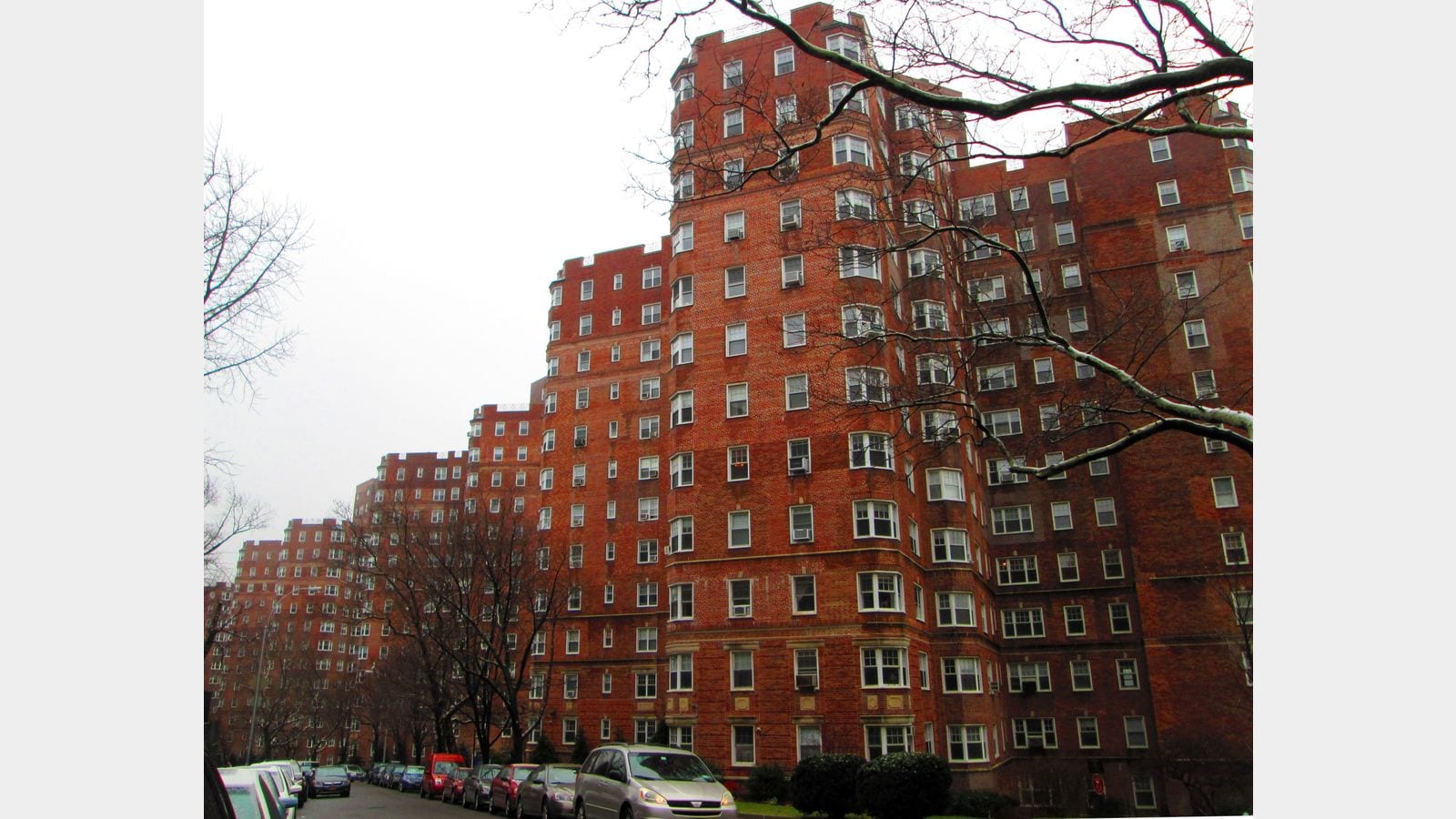Photograph of a row of tall red-brick apartment buildings with bay windows and window air conditioners, stepping down a curving street of parked cars, seen through bare tree branches on a gray day