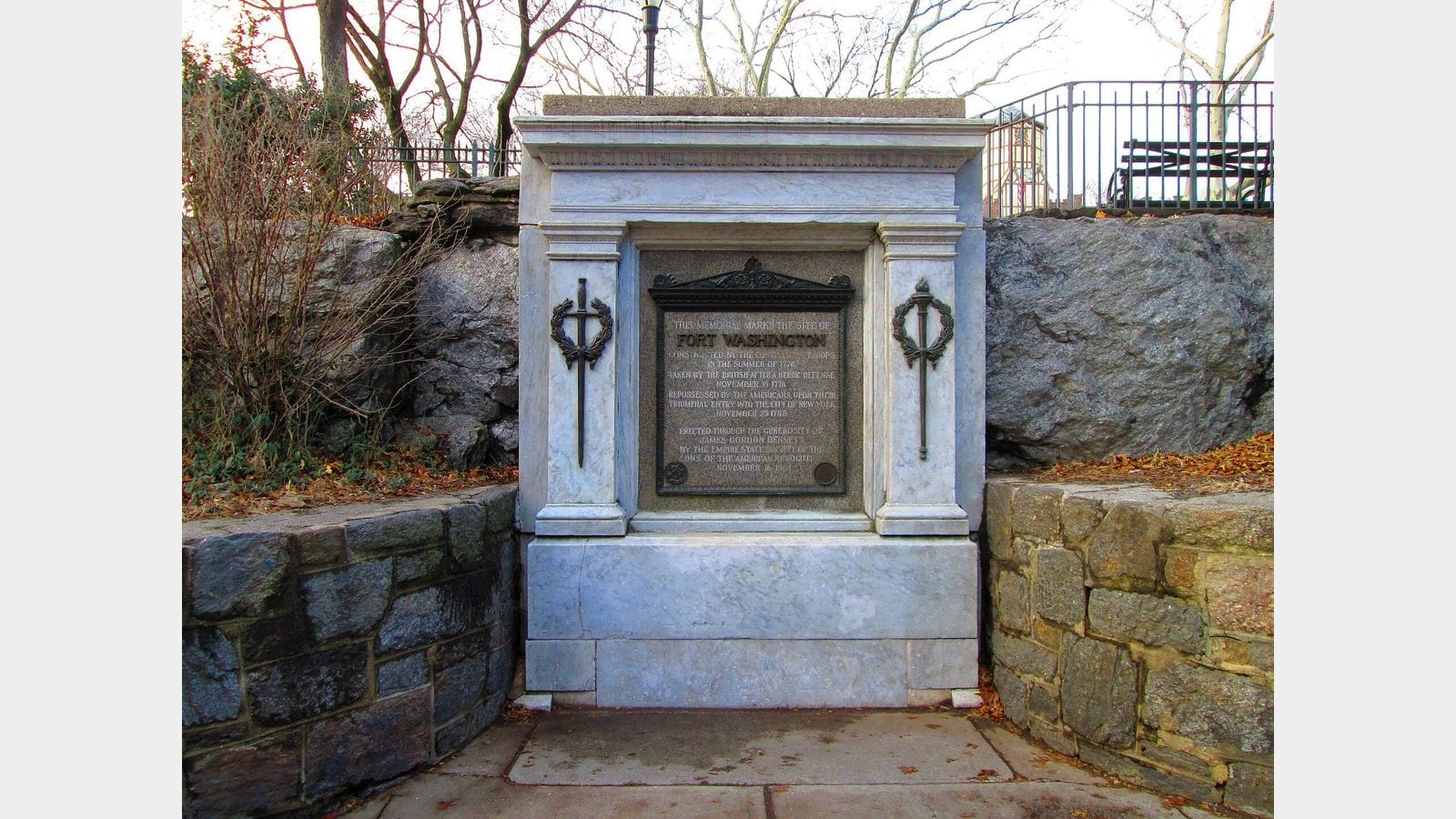 Photograph of a white marble memorial with a bronze plaque reading This memorial marks the site of Fort Washington, set between stone walls and rock outcrops, with bare trees and an iron fence behind