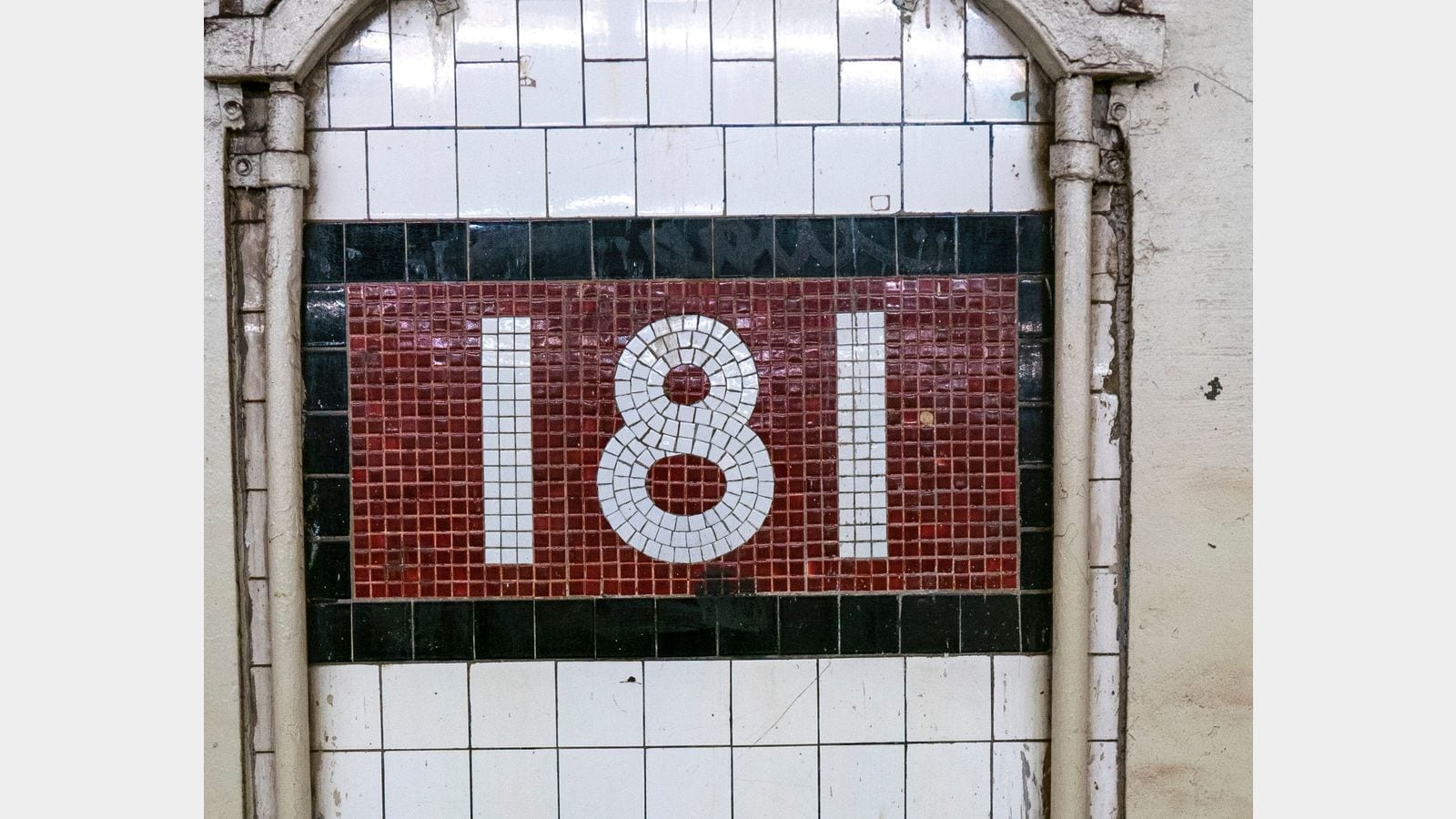 Photograph of a red mosaic tablet reading 181 in white tiles, framed by black tiles and white wall tile under a painted metal arch, in a subway station