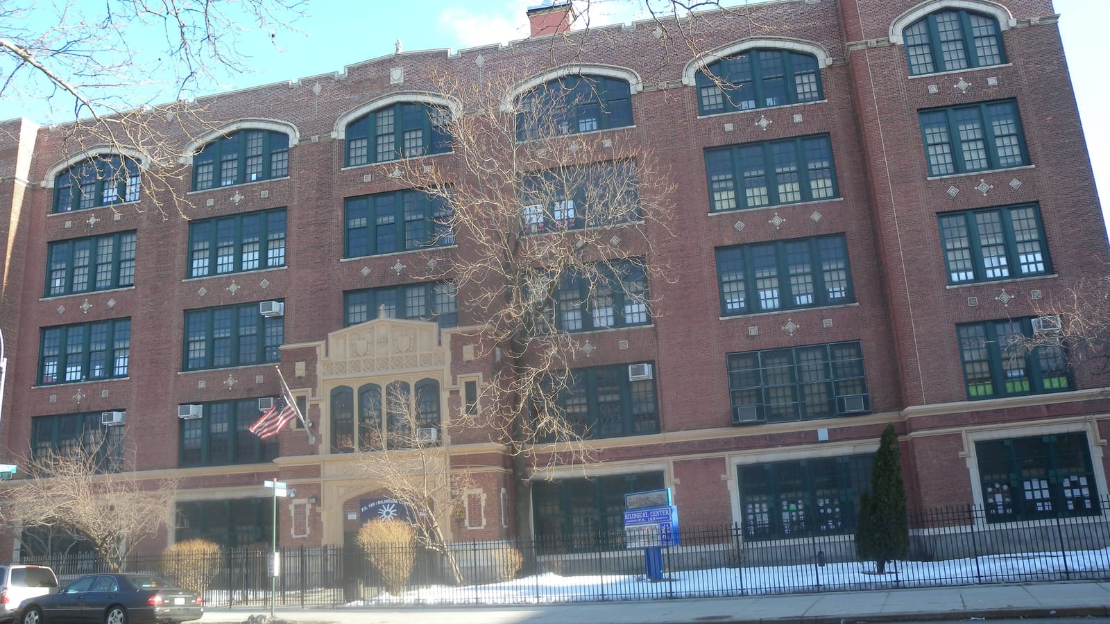 Photograph of a five-story red brick school building with arched upper windows, a carved stone entrance, an American flag, a blue sign reading Bilingual Center P.S. 189, bare trees, a black iron fence, snow on the ground and two parked cars on the street