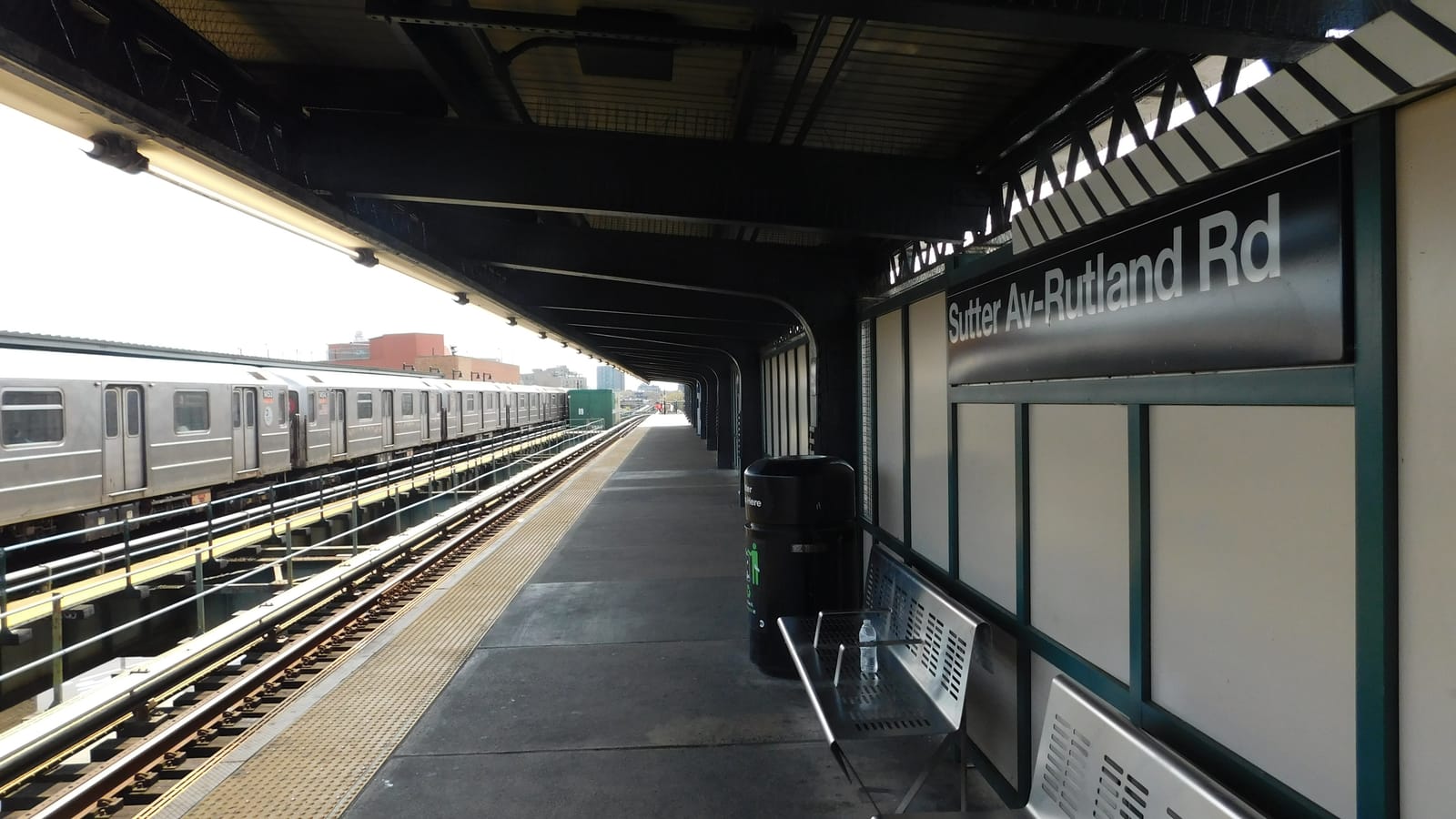 Photograph under the steel canopy of an elevated subway platform, with a black sign reading Sutter Av-Rutland Rd on a white wall at right, two metal benches and a trash can, the platform running into the distance, and a silver subway train on the far track at left