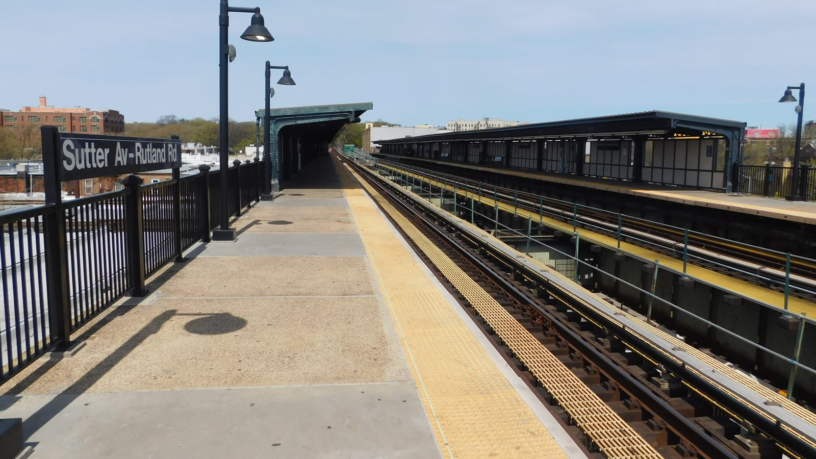 Photograph of an elevated subway platform on a sunny day, with a black sign reading Sutter Av-Rutland Rd on a black railing, black lamp posts, a concrete platform with a yellow edge strip, green-painted canopies over the platforms and tracks to the right, and low brick buildings and trees in the distance