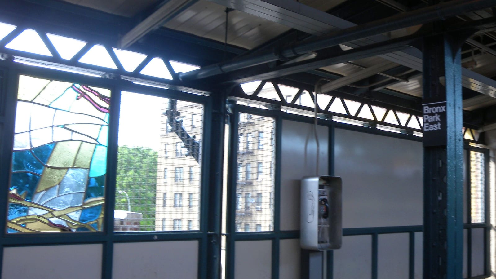 Photograph of an elevated subway platform with a stained-glass panel in blue, white and gold, steel framing, a pay phone on the wall, a black sign reading Bronx Park East on a green column and apartment buildings visible through the windows