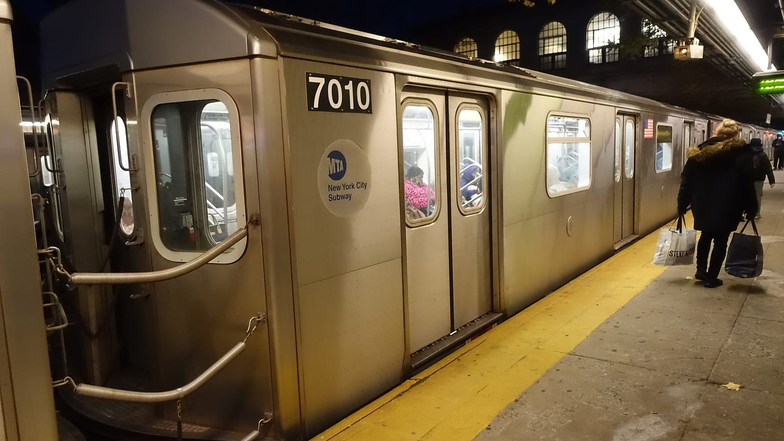 Photograph of a silver subway car numbered 7010 stopped at a platform at night, with its doors closed, a yellow platform edge, a person carrying shopping bags walking away along the platform and arched station windows lit above