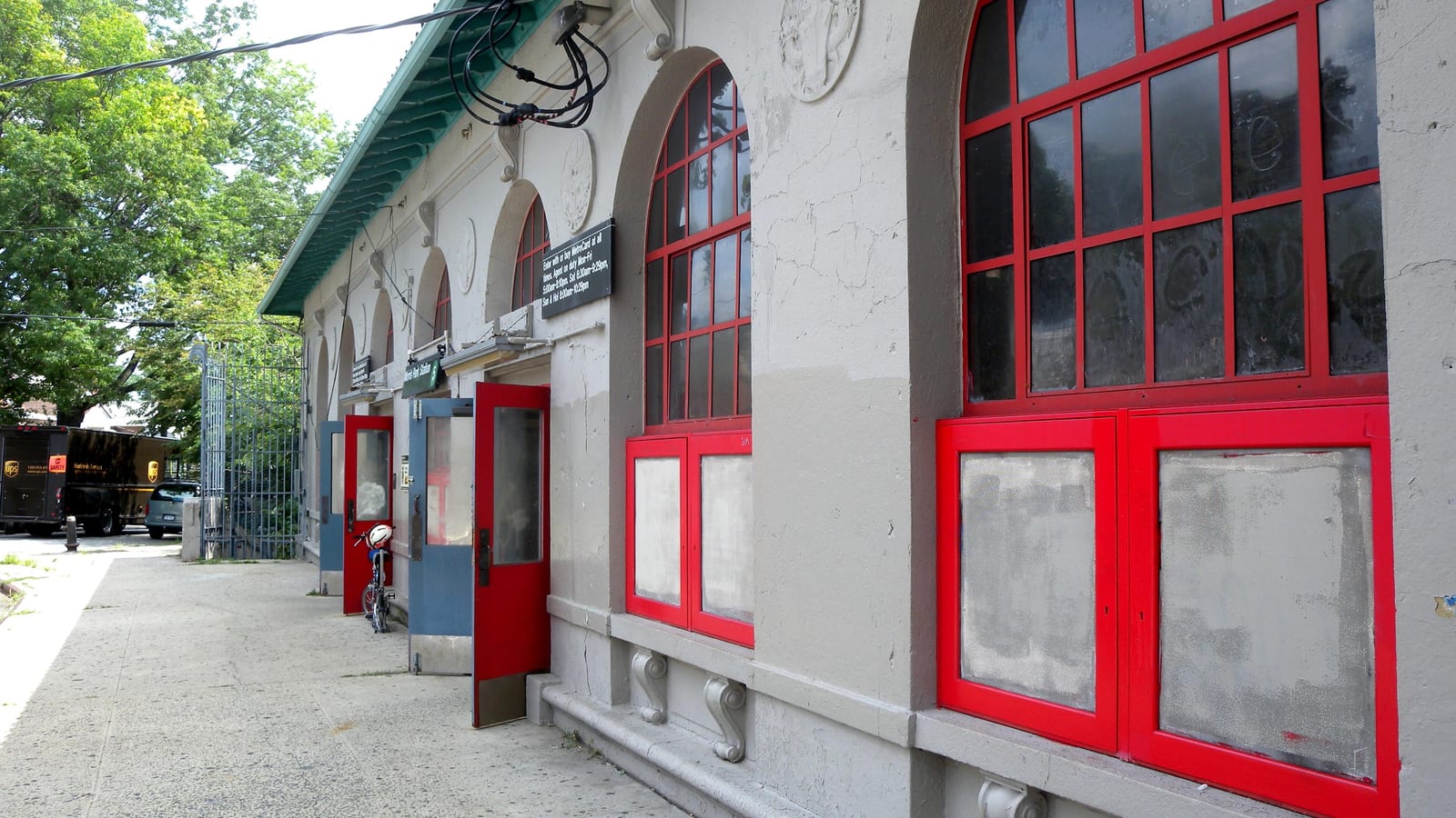 Photograph of the side of a pale stone station building with tall arched windows framed in red, red and blue doors, carved brackets under the window sills, a green tiled roof edge, a wide sidewalk and trees at left under a bright sky