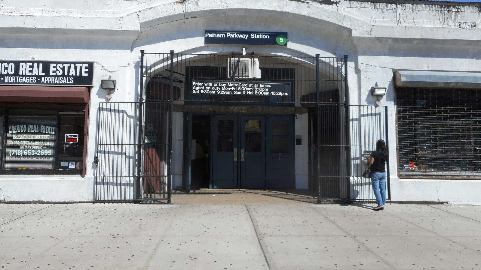 Photograph of a white arched station entrance with a sign reading Pelham Parkway Station and a green 5, open black metal gates, double doors, a posted agent-hours notice, a real estate office at left and a woman standing at the gate seen from the side