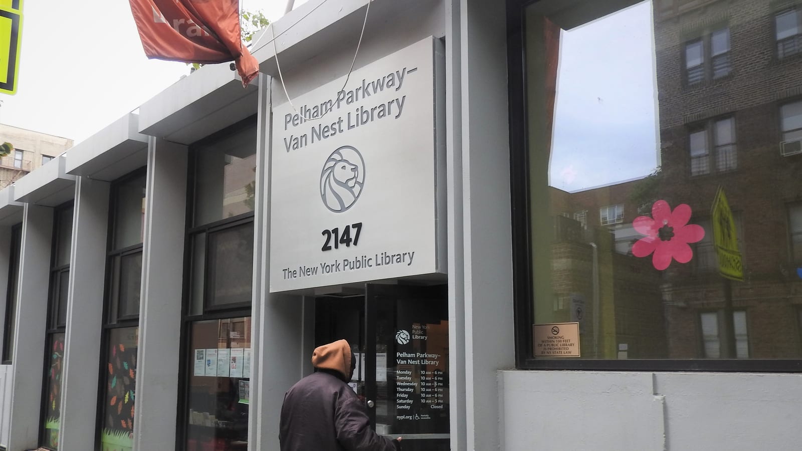 Photograph of a library entrance with a large gray sign reading Pelham Parkway-Van Nest Library, 2147, The New York Public Library, with a lion logo, glass doors with posted hours, a pink flower decal in a window and a person in a hooded jacket at the door seen from behind