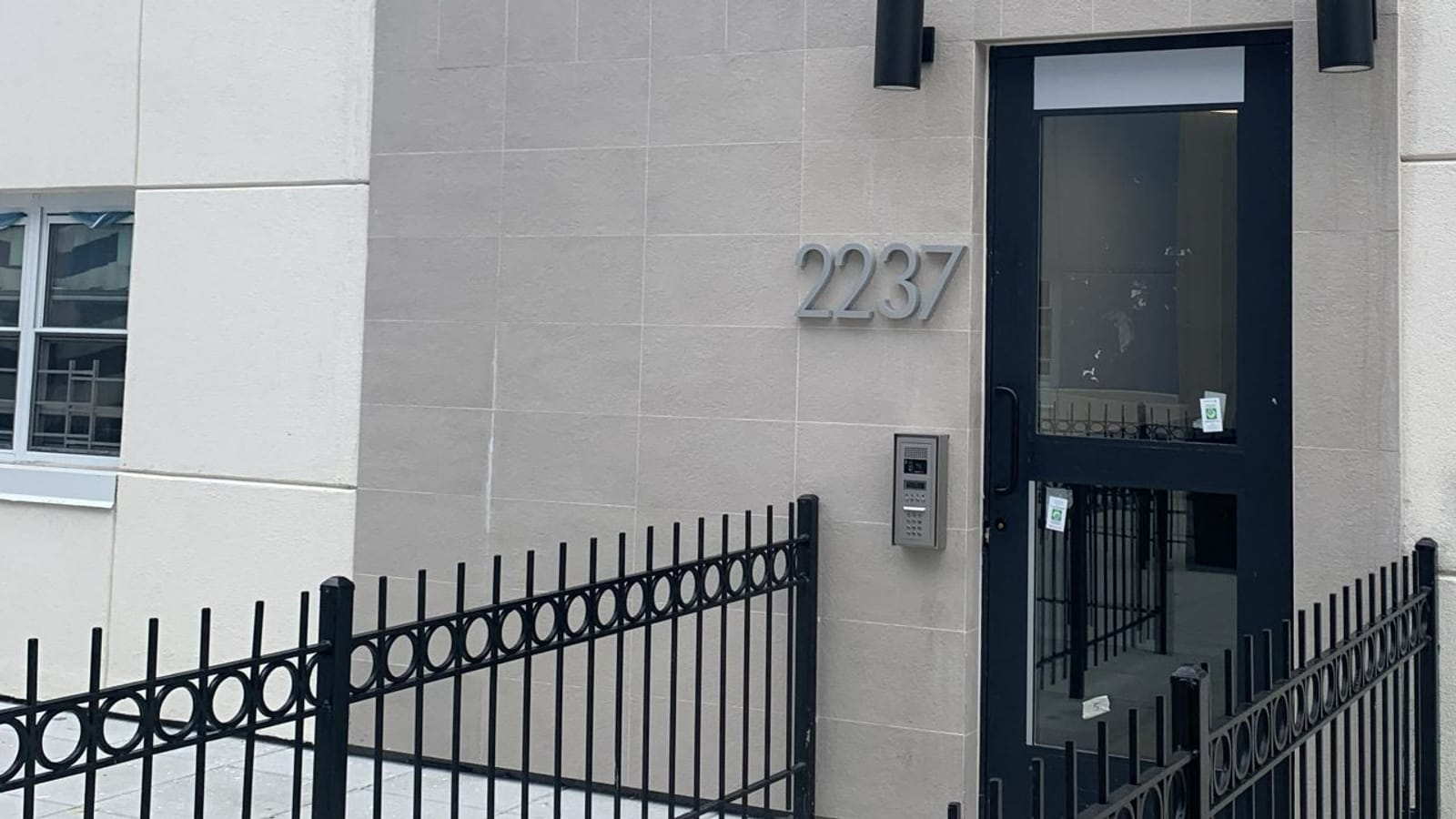 Photograph of a building entrance with a black-framed glass door, the number 2237 in gray metal on a tan stone wall, a keypad intercom panel beside the door, two black wall lights above and a black iron fence in front