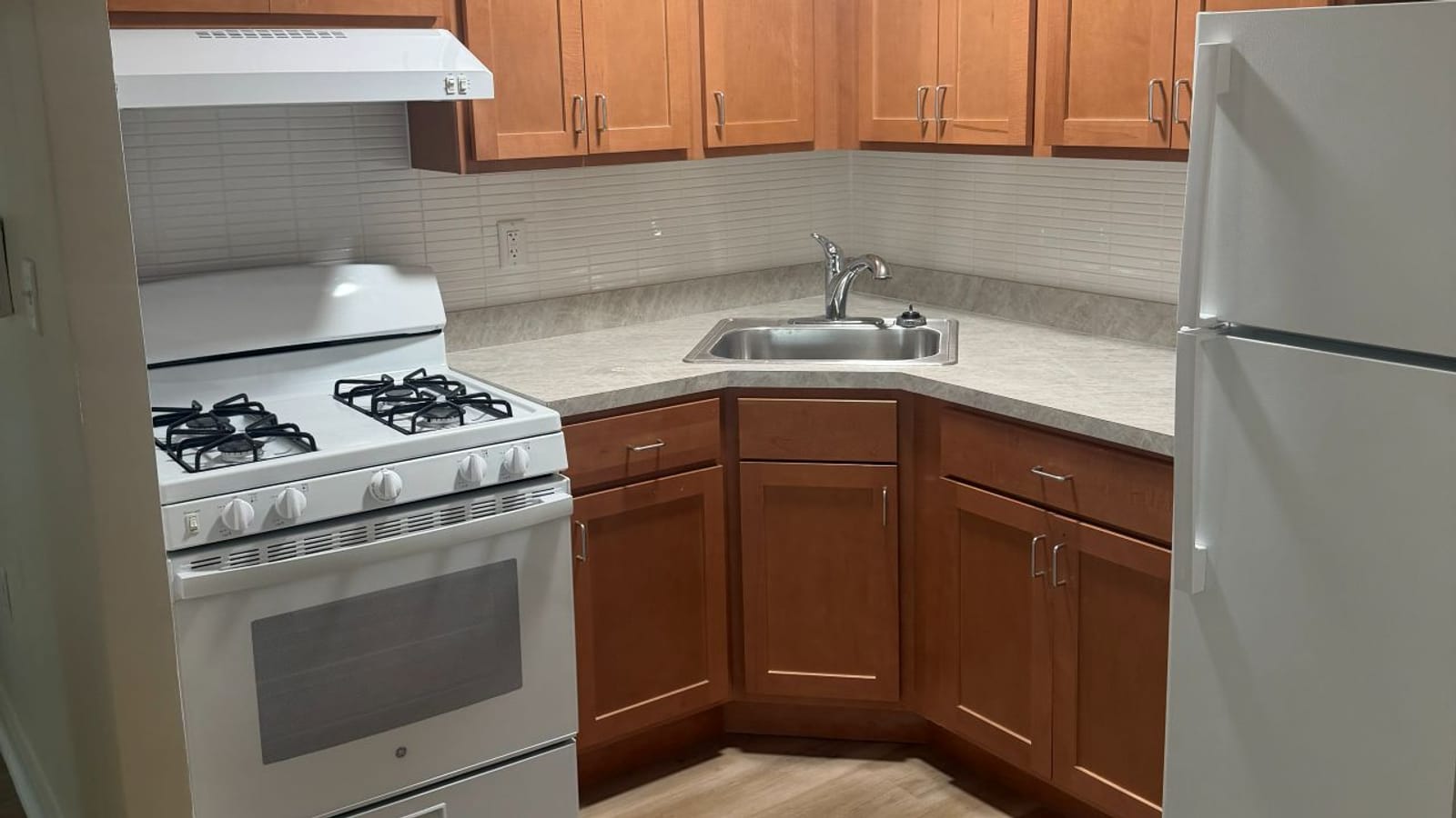 Photograph of a small corner kitchen with wood cabinets, a white gas range under a white hood, a stainless steel sink, a gray counter, a white tiled backsplash and a white refrigerator