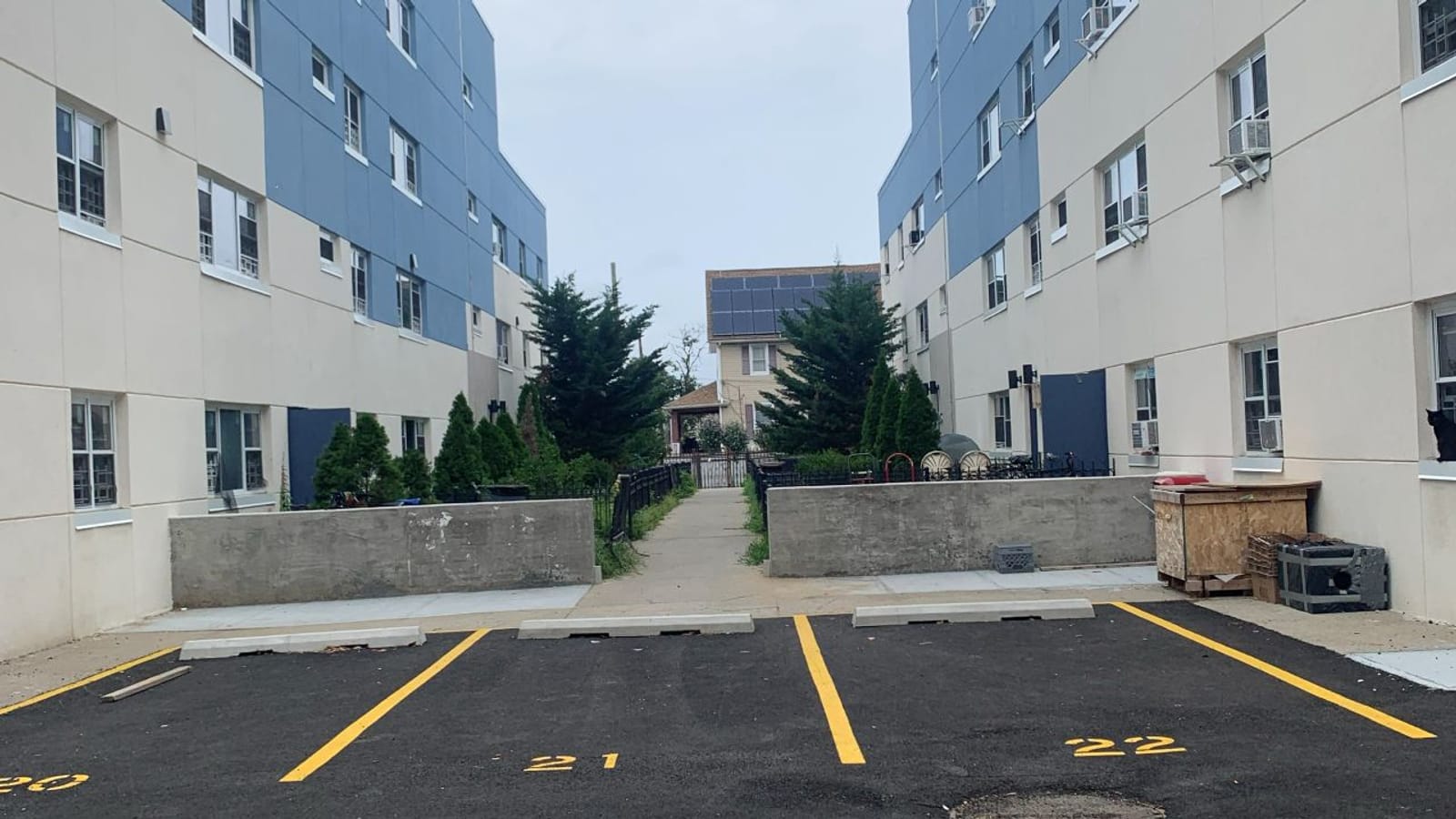 Photograph of a small parking lot with yellow-lined spaces numbered 21 and 22 between two four-story blue and cream apartment buildings, with low concrete walls, evergreen trees and a walkway leading to a house beyond