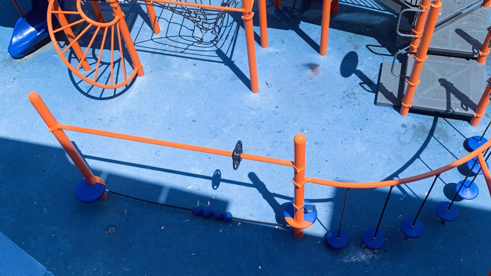 Photograph taken from above of a playground with orange posts, a rope net, a curved climbing frame, blue stepping disks on chains and a blue slide at the top left, on blue rubber ground in sun and shadow