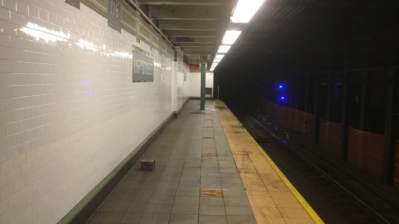 Photograph of an empty underground subway platform with white tiled walls, a mosaic sign reading 116th Street, a green steel column, a yellow platform edge and dark tracks at right