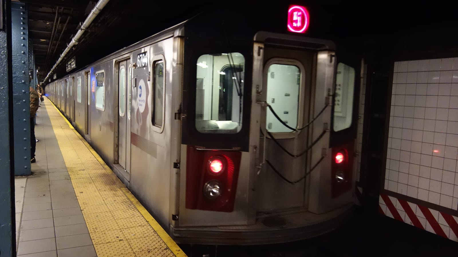 Photograph of a silver subway train with a red 5 sign and red tail lights at an underground platform with blue steel columns, a yellow platform edge and white tiled wall, with two people standing far down the platform