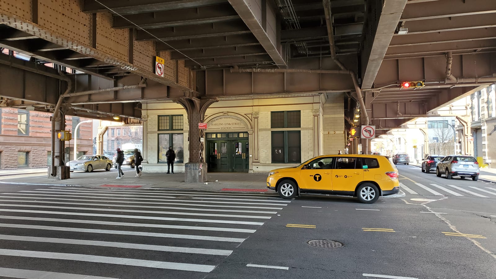 Photograph of a cream stone station entrance with green doors under a dark steel viaduct, a sign over the doors reading Harlem-125th Street Station, a crosswalk and a yellow taxi in the street, and a few people walking on the sidewalk