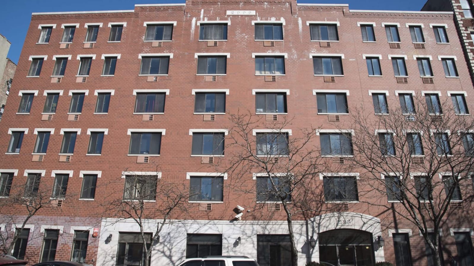 Photograph of a six-story red brick building with rows of white-trimmed windows and wall air conditioners, a stone plaque reading 2000 near the roofline, a pale stone ground floor with an arched entrance marked 223, bare street trees and parked cars in front, under a clear blue sky