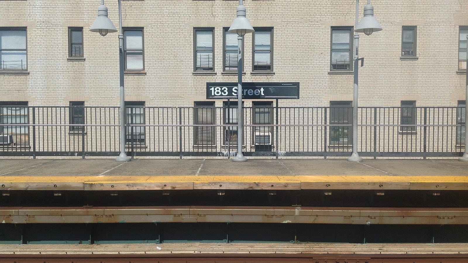 Elevated subway platform with a black 183 Street sign, gray curved lamp posts and a dark railing in front of a tan brick apartment building with window air conditioners, with the yellow platform edge and the tracks in the foreground