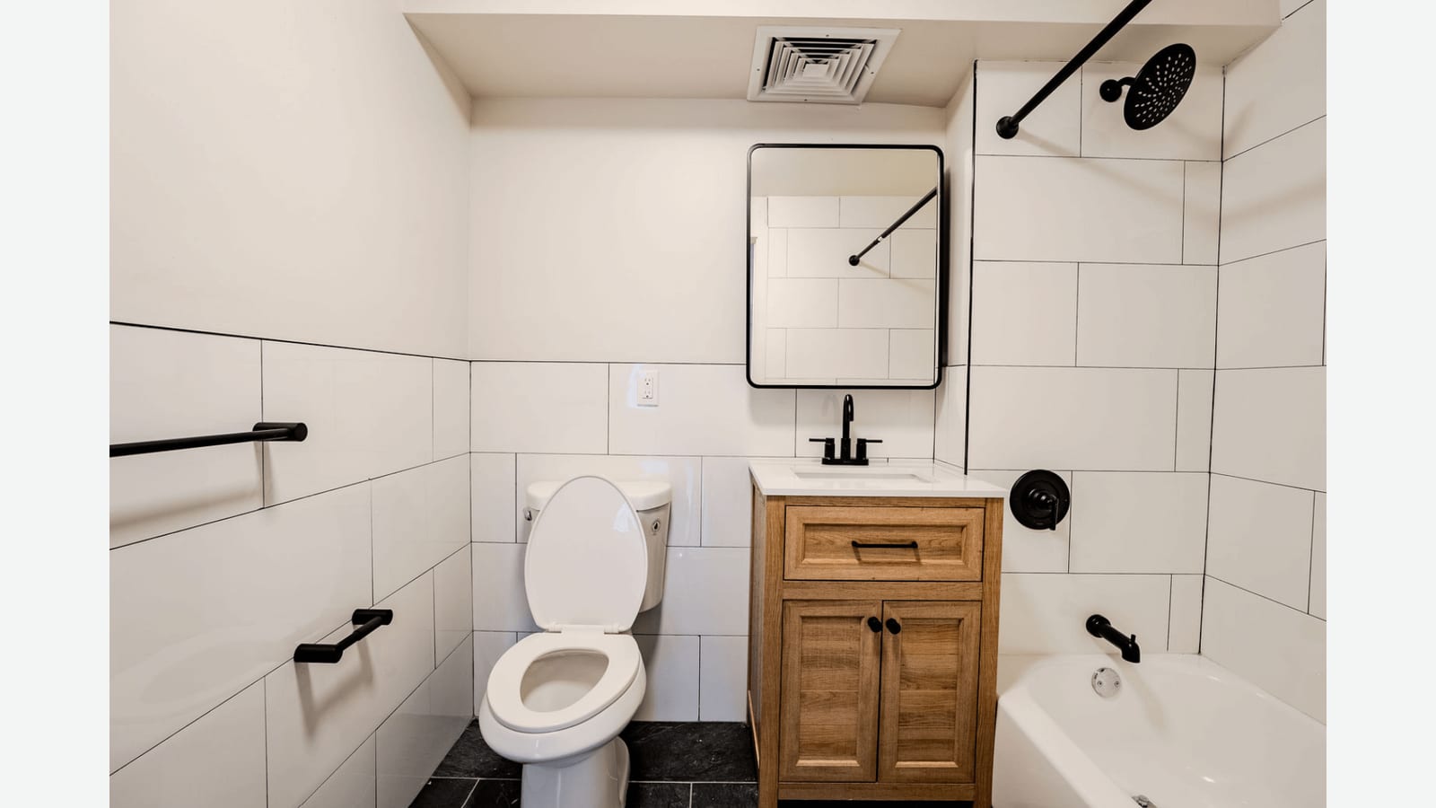Photograph of a bathroom with large white wall tiles, a white toilet, a wood-look vanity with a black faucet under a black-framed mirror, a tub with a black rain shower head and black towel bars, on a dark tile floor