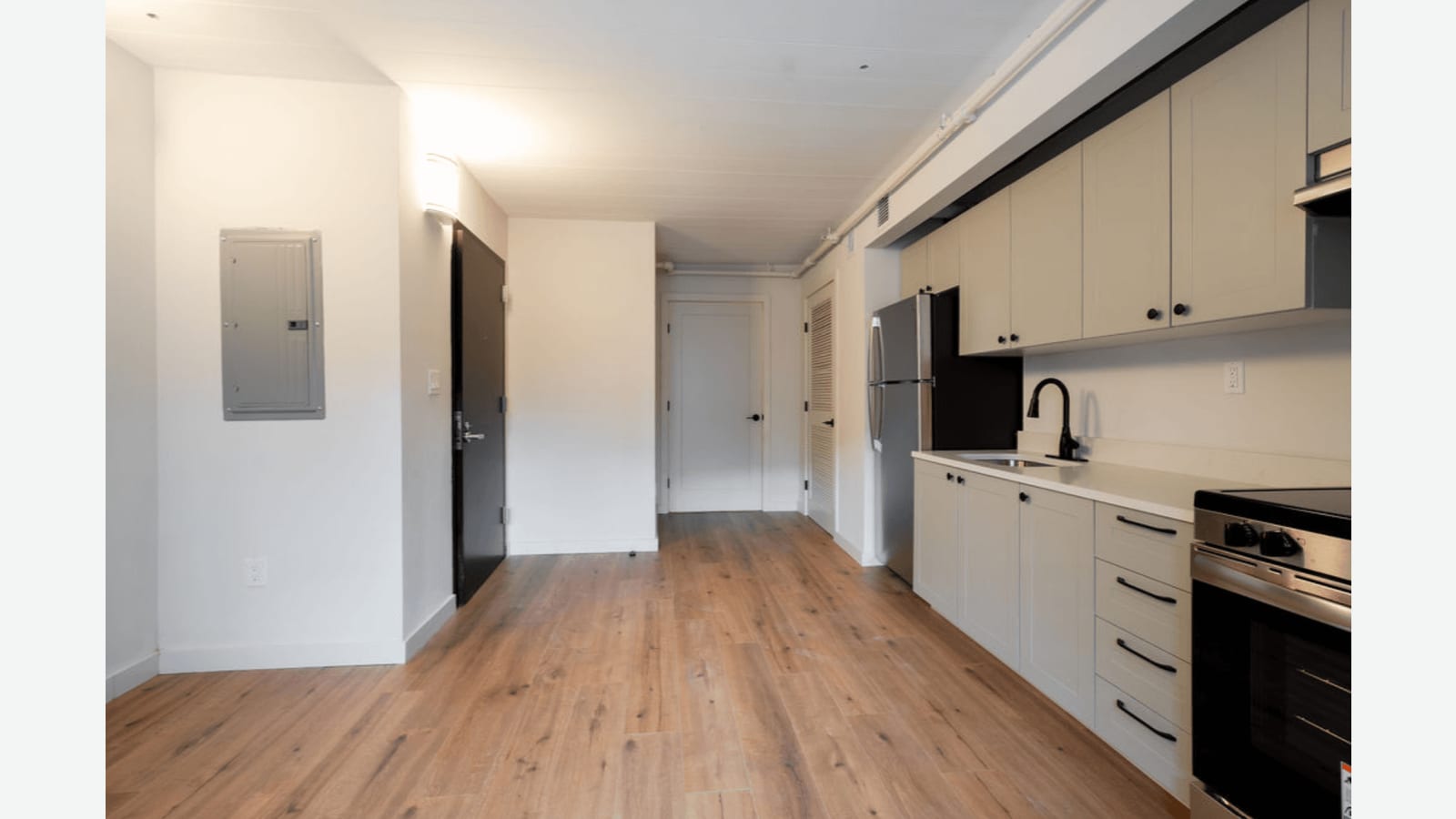 Photograph looking down a long room with a row of light gray kitchen cabinets, a stainless refrigerator and an electric range on the right, a gray electrical panel and a dark entry door on the left, and white doors at the far end
