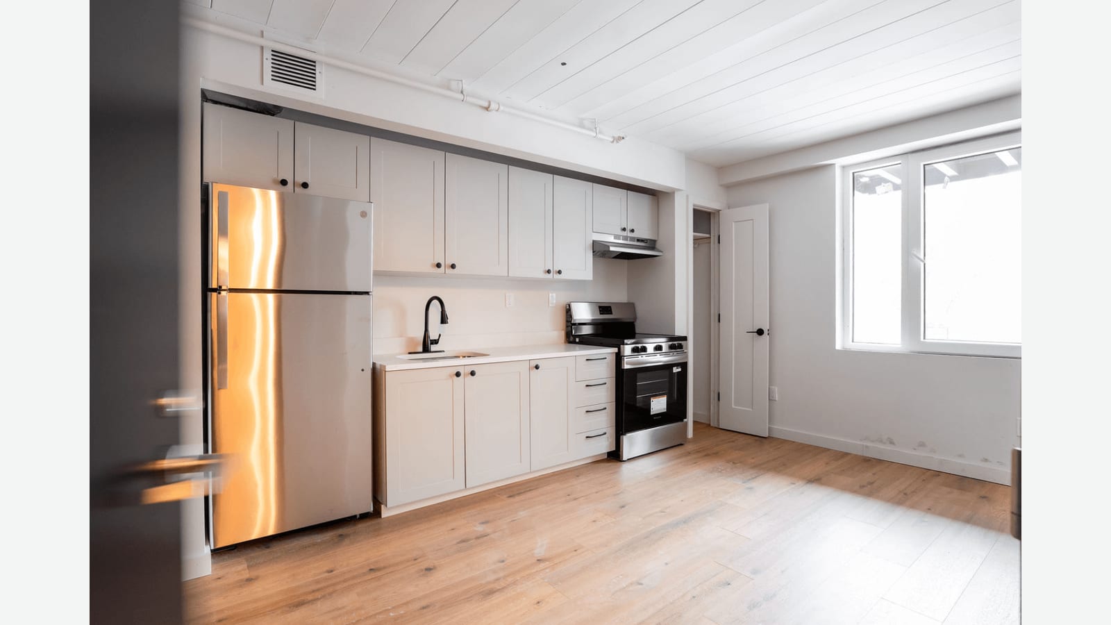 Photograph of a kitchen along one wall with white shaker cabinets, a stainless refrigerator, a white counter with a black faucet, a stainless smooth-top electric range under a hood, white plank ceiling, light wood-look floor and a wide window on the right