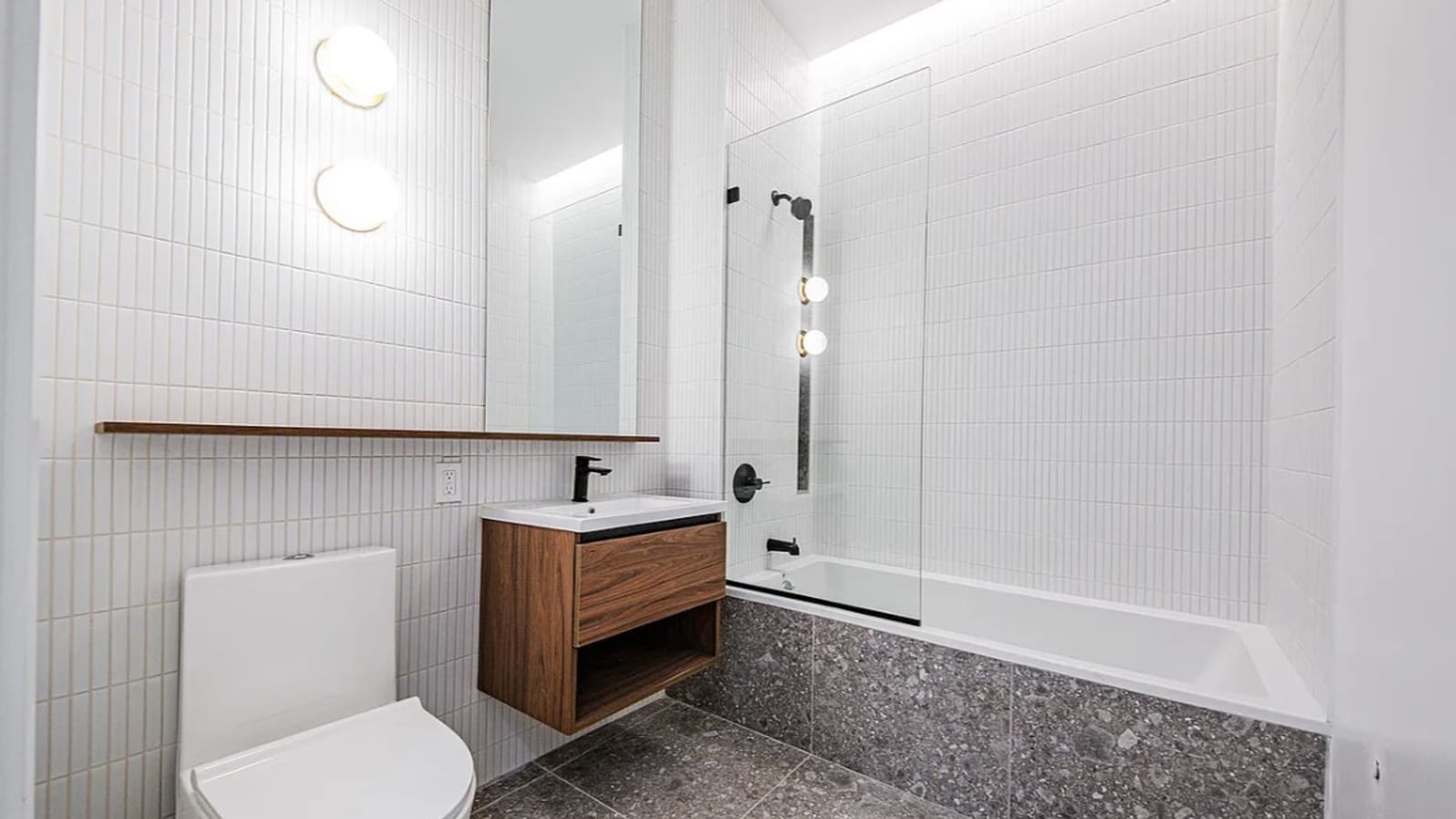 A bathroom with white vertical tile: two round wall lights beside a tall mirror over a narrow wood shelf, a wall-hung walnut vanity with a black faucet, a white toilet, and a bathtub with a glass panel and black shower fixtures, with grey speckled tile on the floor and the front of the tub