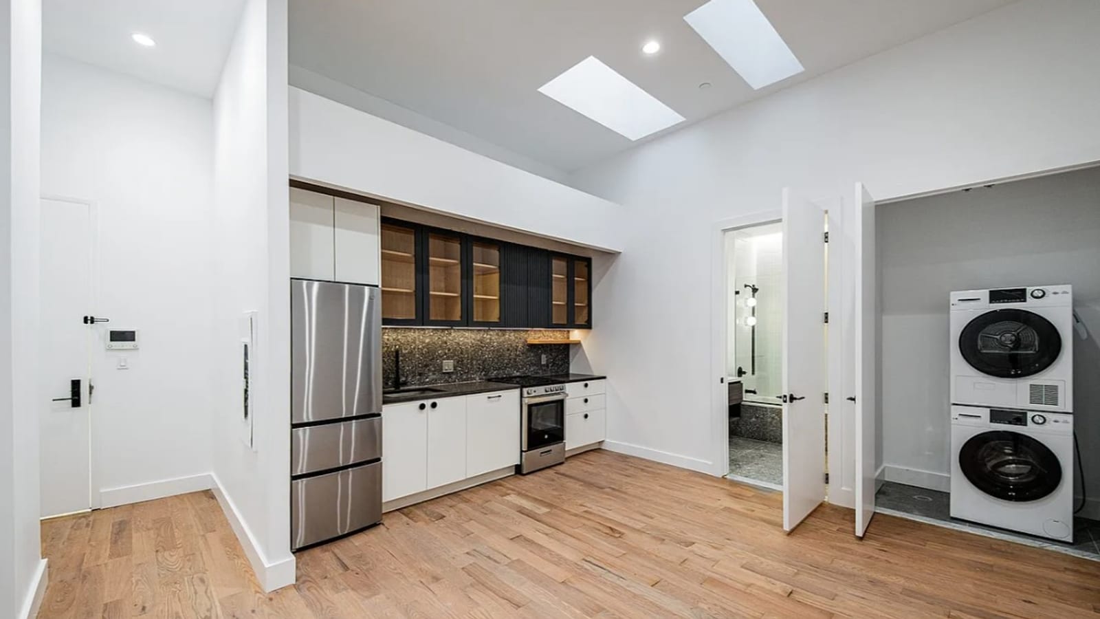 An open kitchen and living space with white walls, two skylights in a high ceiling and light oak floors: a stainless steel refrigerator, white lower cabinets, black glass-front upper cabinets over a dark speckled backsplash, a stainless range, a wall-mounted air conditioner at upper right, an open bathroom door and a closet holding a stacked washer and dryer